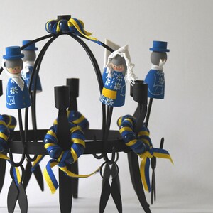 Puede incluir: Un candelabro de metal negro con m&uacute;ltiples portavelas. Est&aacute; decorado con cintas azules y amarillas y peque&ntilde;as figuras de madera con trajes tradicionales suecos. Las figuras est&aacute;n pintadas con patrones florales azules y blancos.