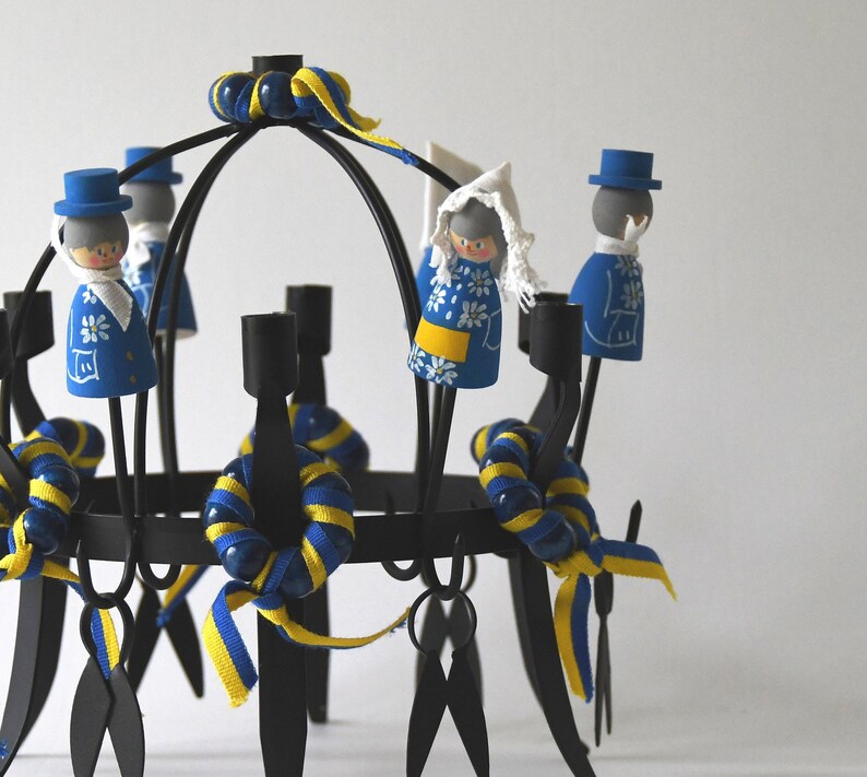 Puede incluir: Un candelabro de metal negro con m&uacute;ltiples portavelas. Est&aacute; decorado con cintas azules y amarillas y peque&ntilde;as figuras de madera con trajes tradicionales suecos. Las figuras est&aacute;n pintadas con patrones florales azules y blancos.