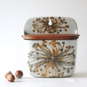 Può includere: Un contenitore in ceramica da parete con coperchio in legno. Il contenitore è bianco con un motivo floreale marrone e giallo. Tre nocciole sono davanti al contenitore.
