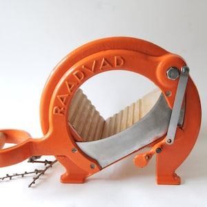 Puede incluir: Cortadora de pan RAADVAAD naranja con mango de madera y cuchilla de acero inoxidable. La cortadora tiene un diseño circular con el nombre de la marca en relieve.