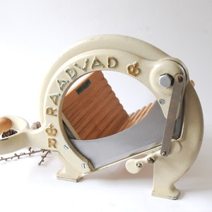 Puede incluir: Cortador de pan de hierro fundido blanco con una guía de pan de madera y una cuchilla plateada. El cortador está etiquetado "R & B RADVAD &"