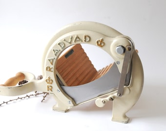 May include: White cast iron bread slicer with a wooden bread guide and a silver blade. The slicer is labelled "R & B RADVAD &"