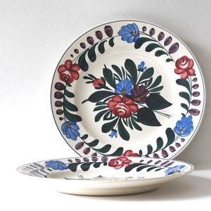 May include: Two white ceramic plates with floral designs. The plates feature a central bouquet of red, blue, and purple flowers with green leaves. A border of blue, red, and purple flowers and leaves encircles the plates.