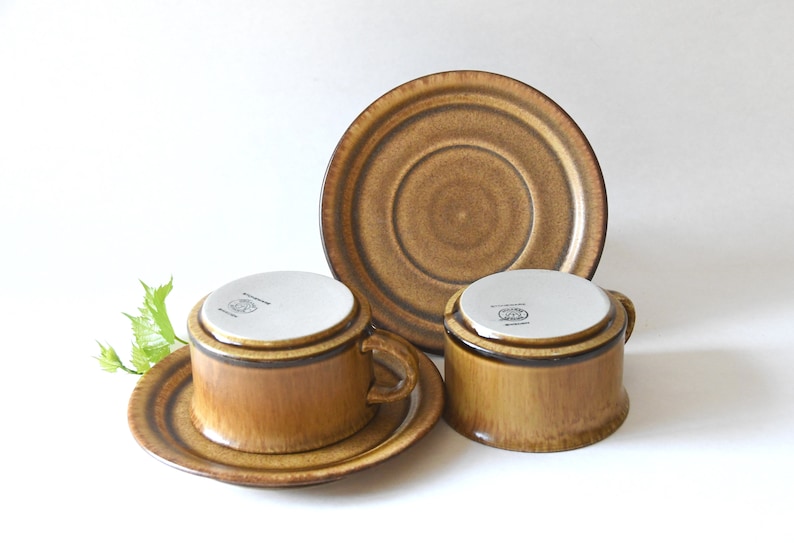 May include: Two brown ceramic coffee cups with matching saucers. The cups have a brown and white glaze and a dark brown rim. The saucers have a brown and white glaze with a dark brown rim.
