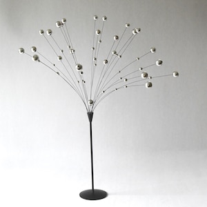 Op de afbeelding: Een decoratief sculptuur met een dunne zwarte metalen standaard en voet die een cluster van uitstralende draadstelen ondersteunt. Elke draadsteel is bekroond met een kleine, gepolijste zilverkleurige bol, wat een waaiervormig arrangement vormt.