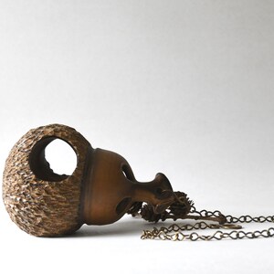 May include: A brown, acorn-shaped pendant with a textured cap and a smooth, hollowed-out base featuring circular openings. The pendant is attached to a metallic chain that includes small, decorative pinecone-like charms near the connection point.