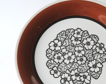 以下が含まれることがあります： 茶色の縁取りと白黒の花柄が描かれた白いセラミック製の皿。花は様式化されており、ビンテージな雰囲気です。