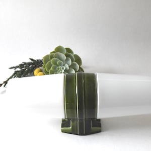 Puede incluir: Jarrón de cerámica blanco con base verde esmaltada. El jarrón está decorado con una serie de rayas horizontales verdes. Una planta suculenta se coloca en el jarrón.