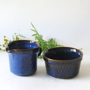 Puede incluir: Dos piezas de vajilla de cerámica azul. Una es una jarra pequeña con asa y boquilla, la otra es un cuenco redondo. Ambos tienen una superficie texturizada y un borde marrón. El fondo es blanco con algo de vegetación.