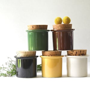 Può includere: Una collezione di piccoli vasetti in ceramica di vari colori, tra cui verde, marrone, nero, giallo e bianco. Ogni vasetto è sormontato da un tappo in sughero naturale. Due pompon gialli sono posti sopra un vasetto. I vasetti sono disposti a piramide.