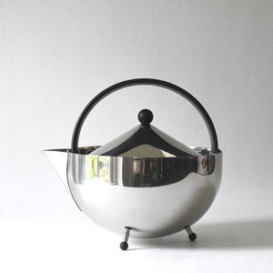 Puede incluir: Una tetera de plata con asa y tapa negras. La tetera tiene un cuerpo redondo y tres patas.