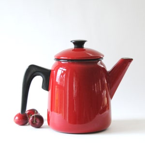 May include: A red enamel teapot with a black handle and lid. The teapot is sitting on a white surface with three red cherries in front of it.