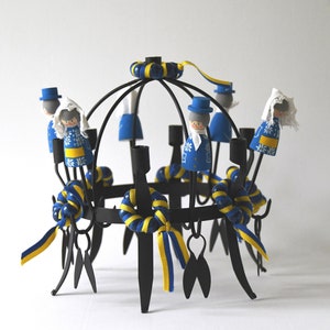 Puede incluir: Un candelabro de metal negro con un dise&ntilde;o circular, adornado con cintas azules y amarillas y peque&ntilde;as figuras de madera vestidas con trajes tradicionales. Las figuras llevan abrigos azules y gorros blancos. El candelabro mide unos 25 cm de alto.