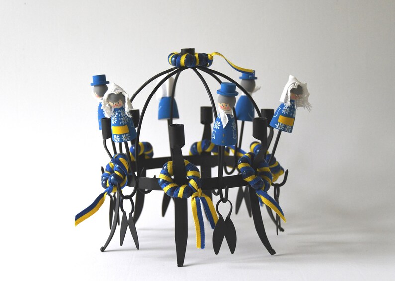 Puede incluir: Un candelabro de metal negro con un dise&ntilde;o circular, adornado con cintas azules y amarillas y peque&ntilde;as figuras de madera vestidas con trajes tradicionales. Las figuras llevan abrigos azules y gorros blancos. El candelabro mide unos 25 cm de alto.
