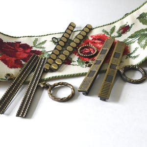Könnte beinhalten: Eine Auswahl an antiken Messingbeschlägen, darunter Schubladengriffe und Ringgriffe, angeordnet mit einem floralen bestickten Band mit roten Rosen und grünen Blättern. Die Hardware-Teile haben verschiedene strukturierte Designs.