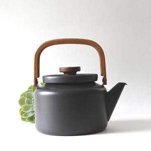 Puede incluir: Una tetera de cerámica gris oscuro con asa y pomo de madera. La tetera tiene un pico y se apoya sobre una superficie blanca. Una planta suculenta verde está al lado de la tetera.