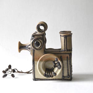 Pode incluir: Uma escultura de cerâmica bege em forma de câmara de cinema vintage com detalhes esmaltados em castanho escuro. A peça apresenta uma lente circular, uma pega superior e um elemento vertical, decorado com padrões lineares e pontos escuros.