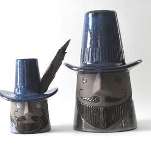 Op de afbeelding: Twee keramische sculpturen van grillige figuren met hoge hoeden en snorren. Eén figuur heeft een veer in zijn hoed. De figuren zijn voornamelijk blauw en bruin, met een glanzende afwerking. De sculpturen hebben een unieke uitstraling.
