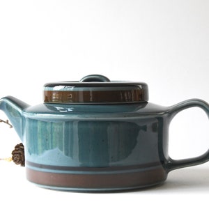 May include: A ceramic teapot featuring a glossy teal blue glaze with a dark brown band around the base and a matching lid. The teapot has a short spout, a curved handle, and a flat, cylindrical body shape with a smooth, reflective finish.