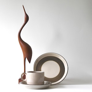 May include: A wooden crane sculpture stands tall next to a beige teacup and saucer, and a plate with brown and beige concentric circles. The items are arranged on a white surface, creating a minimalist aesthetic.