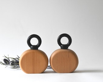 May include: Two wooden, circular objects with a light brown finish. Each has a black ring on top. The objects are displayed on a white background. A sprig of dried, blue-green flowers is to the left.