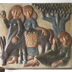 Könnte beinhalten: Eine quadratische Keramik-Wandfliese mit einem Relief von zwei stehenden und einer liegenden Figur sowie einem Esel, der einen Korb mit Früchten trägt. Die Szene zeigt einen Baum im Hintergrund in erdigen Braun-, Blau- und Beigetönen.