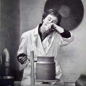 May include: A woman in a white coat is working on a pottery wheel. She is holding a piece of clay in her hands and is concentrating on her work. The pottery wheel is made of wood and has a metal base. The woman is wearing a necklace and has her hair pulled back.