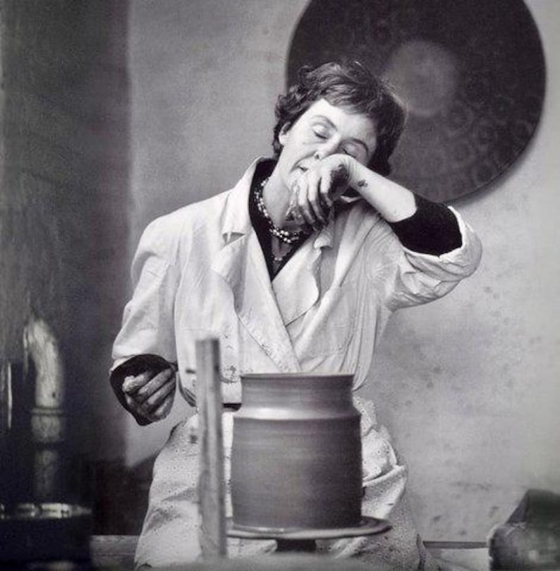 May include: A woman in a white coat is working on a pottery wheel. She is holding a piece of clay in her hands and is concentrating on her work. The pottery wheel is made of wood and has a metal base. The woman is wearing a necklace and has her hair pulled back.