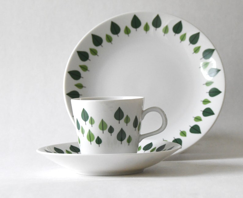 Puede incluir: Un juego de vajilla de cer&aacute;mica blanca con una taza, un platillo y un plato llano. Cada pieza est&aacute; decorada con un patr&oacute;n de hojas verdes. El plato y la taza tienen un borde de hojas, mientras que la taza y el platillo tienen un dise&ntilde;o de hojas dispersas.