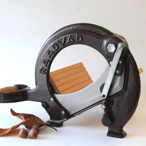 Puede incluir: Un cascanueces vintage de hierro fundido con una tabla de cortar de madera y una cuchilla plateada. El cascanueces está etiquetado con "RAADVAAD" en el lateral.