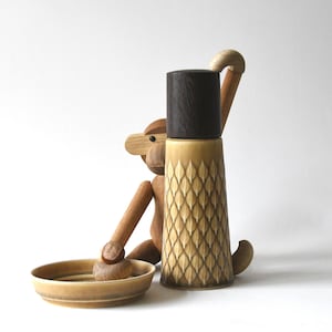 Puede incluir: Un molinillo de pimienta de cerámica beige con una tapa marrón oscuro y un patrón de diamantes. Una figura de mono de madera está sentada junto al molinillo, con un cuenco pequeño y un mortero de madera.