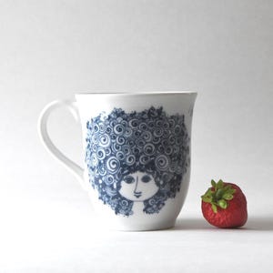 Può includere: Tazza in ceramica bianca con un'illustrazione blu e bianca del volto di una donna con i capelli ricci. Una fragola rossa con foglie verdi è accanto alla tazza.