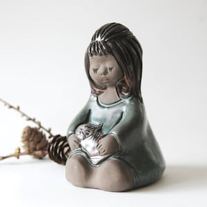 May include: A ceramic figurine of a girl with long dark hair, wearing a teal blue dress and holding a small animal in her arms. The figurine has a rustic, hand-crafted look.