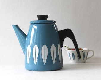 May include: A blue enamel teapot with a black handle and lid knob, featuring a white leaf-like pattern around the base. Beside it sits a small white creamer with a matching blue leaf pattern, containing a few small red cherries inside.