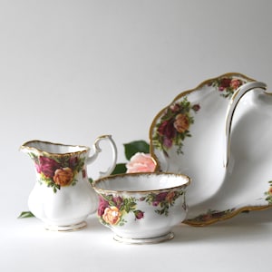 以下が含まれることがあります： 白磁のティーセット。クリーマー、砂糖入れ、サービング皿が含まれています。各ピースは、赤、黄色、緑の色合いのバラのパターンで装飾され、金色のトリムが施されています。ピンクのバラが近くに置かれています。