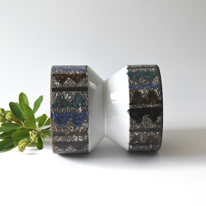 May include: A white ceramic vase with an unusual hourglass form. The ends of the vase feature a textured pattern in shades of brown, blue, and black. A sprig of green leaves and buds sits beside the vase, adding a natural touch.