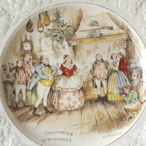 May include: A cream-coloured ceramic plate with a raised floral border and a central illustration of a group of people in period clothing dancing in a room. The text "CHRISTMAS EVE AT MR WARDLE'S" and "A.L. STANMORE" are printed at the bottom.