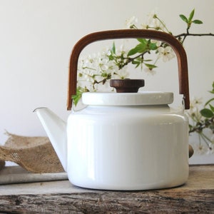 Puede incluir: Una tetera de esmalte blanco con asa y pomo de madera. La tetera tiene un pico curvo y un cuerpo redondeado. El asa es de madera oscura y se arquea sobre la parte superior. El pomo de la tapa también es de madera oscura.