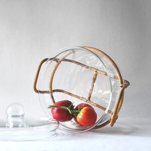 May include: A clear glass serving bowl with a removable rattan frame and two side handles. The bowl is tilted on its side and contains a cluster of three fresh red tomatoes on a green vine. A separate clear glass lid with a knob handle sits nearby.