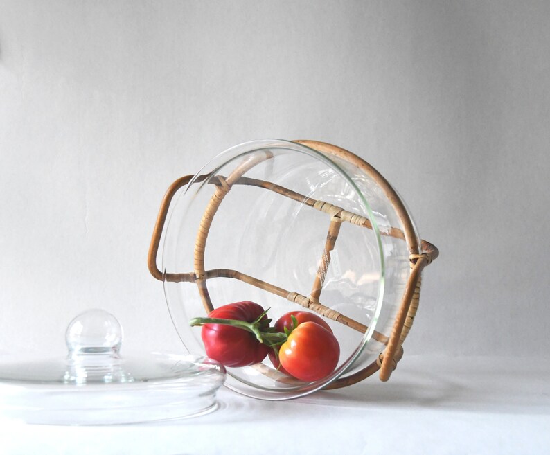 May include: A clear glass serving bowl with a removable rattan frame and two side handles. The bowl is tilted on its side and contains a cluster of three fresh red tomatoes on a green vine. A separate clear glass lid with a knob handle sits nearby.
