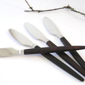 Puede incluir: Cuatro cuchillos de mantequilla de acero inoxidable con mangos de madera marrón oscuro. Los cuchillos están dispuestos en forma de abanico con los mangos superpuestos.