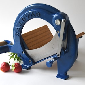 Puede incluir: Un cortador de queso RADVAD de hierro fundido azul con una tabla de cortar de madera y una cuchilla plateada. Dos fresas rojas están en primer plano.