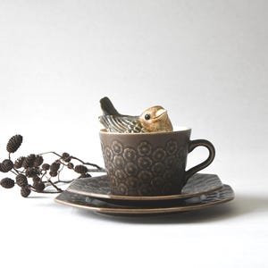 Może przedstawiać: Brązowy ceramiczny zestaw do herbaty z filiżanką i spodkiem z dekoracyjnym kwiatowym wzorem. Mała figurka ptaka znajduje się w filiżance. Gałązka suszonych liści jest umieszczona z boku. Uroczy prezent.