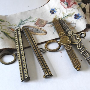 May include: Vintage brass sewing scissors and two measuring tools. The scissors have a heart-shaped detail. The measuring tools have a series of holes. A floral embroidered fabric is in the background. The background is white.