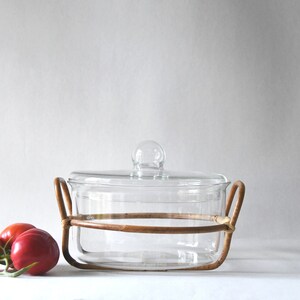 May include: A clear glass casserole dish with a matching glass lid featuring a round knob handle. The dish is cradled in a circular light brown rattan frame with two side handles. Two red tomatoes with green stems are positioned to the left side.