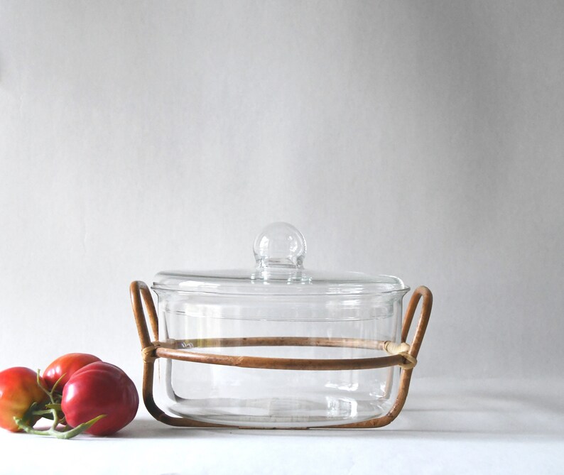 May include: A clear glass casserole dish with a matching glass lid featuring a round knob handle. The dish is cradled in a circular light brown rattan frame with two side handles. Two red tomatoes with green stems are positioned to the left side.