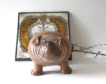 May include: A brown ceramic bulldog figurine with a rounded body, short legs, and incised facial features. Behind the figurine sits a square decorative tile featuring a stylized illustration of a cat with a large, radiating mane.