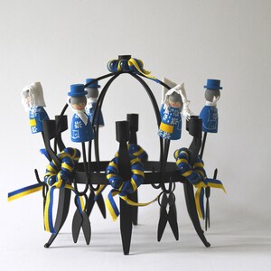 Puede incluir: Un candelabro de metal negro con ocho portavelas. El candelabro est&aacute; decorado con cintas azules y amarillas y peque&ntilde;as figuras de madera vestidas con trajes azules y blancos. Las figuras est&aacute;n dispuestas alrededor de los portavelas.