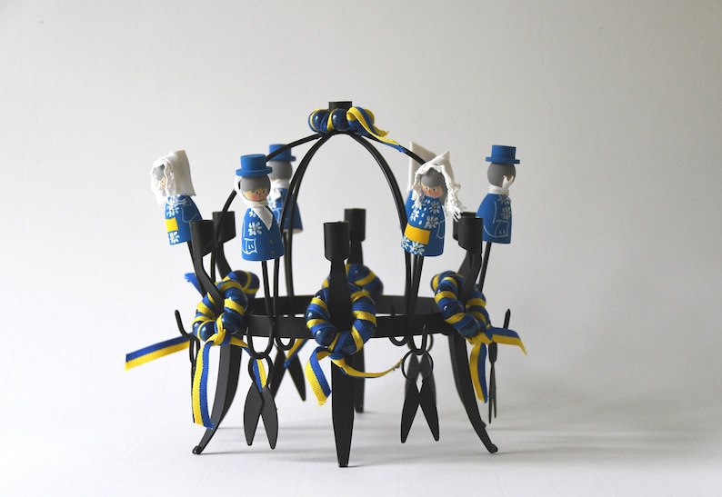 Puede incluir: Un candelabro de metal negro con ocho portavelas. El candelabro est&aacute; decorado con cintas azules y amarillas y peque&ntilde;as figuras de madera vestidas con trajes azules y blancos. Las figuras est&aacute;n dispuestas alrededor de los portavelas.