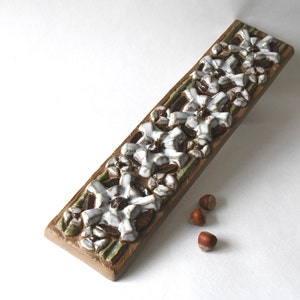 May include: A long, rectangular ceramic tile with a raised design of white flowers on a brown and green background.